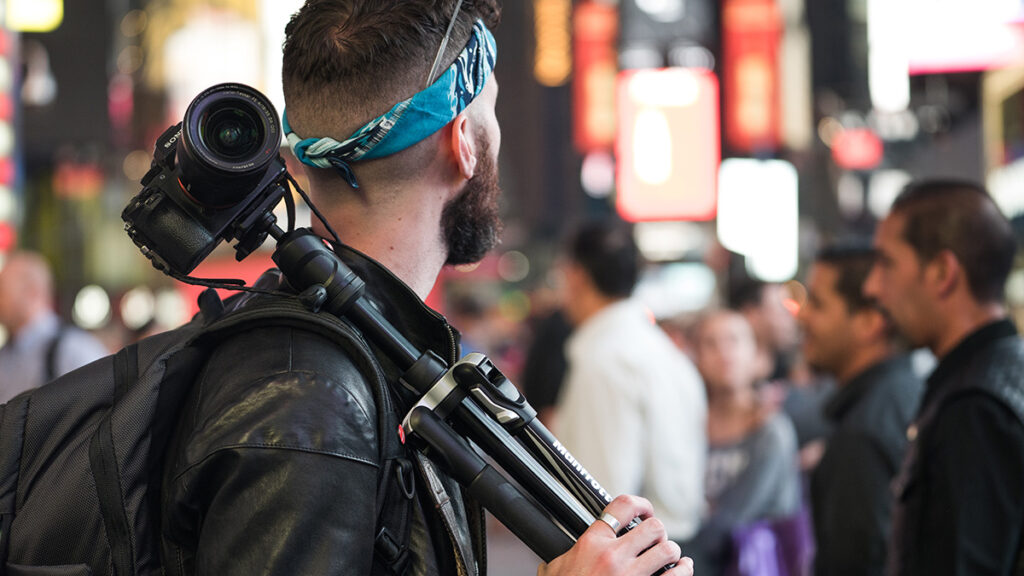 A Photographer holding a tripod with a camera mounted on it and a camera bag hanging over his shoulder.